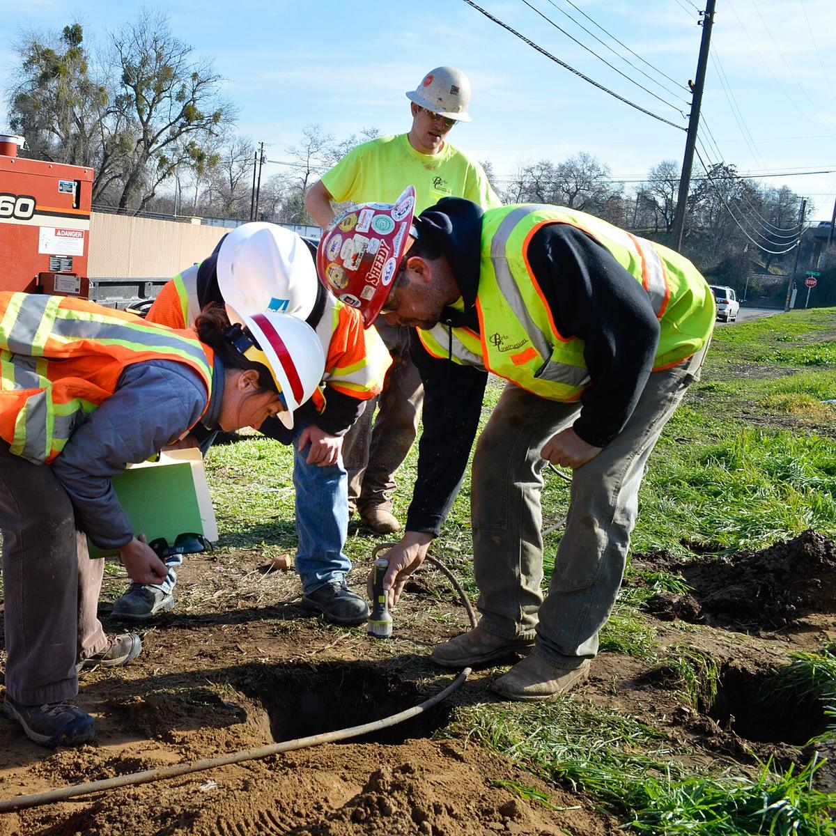 A crew in hard hats and safety vests leans over a small open pothole, looking for a buried gas line, with a vacuum excavation unit parked behind them.