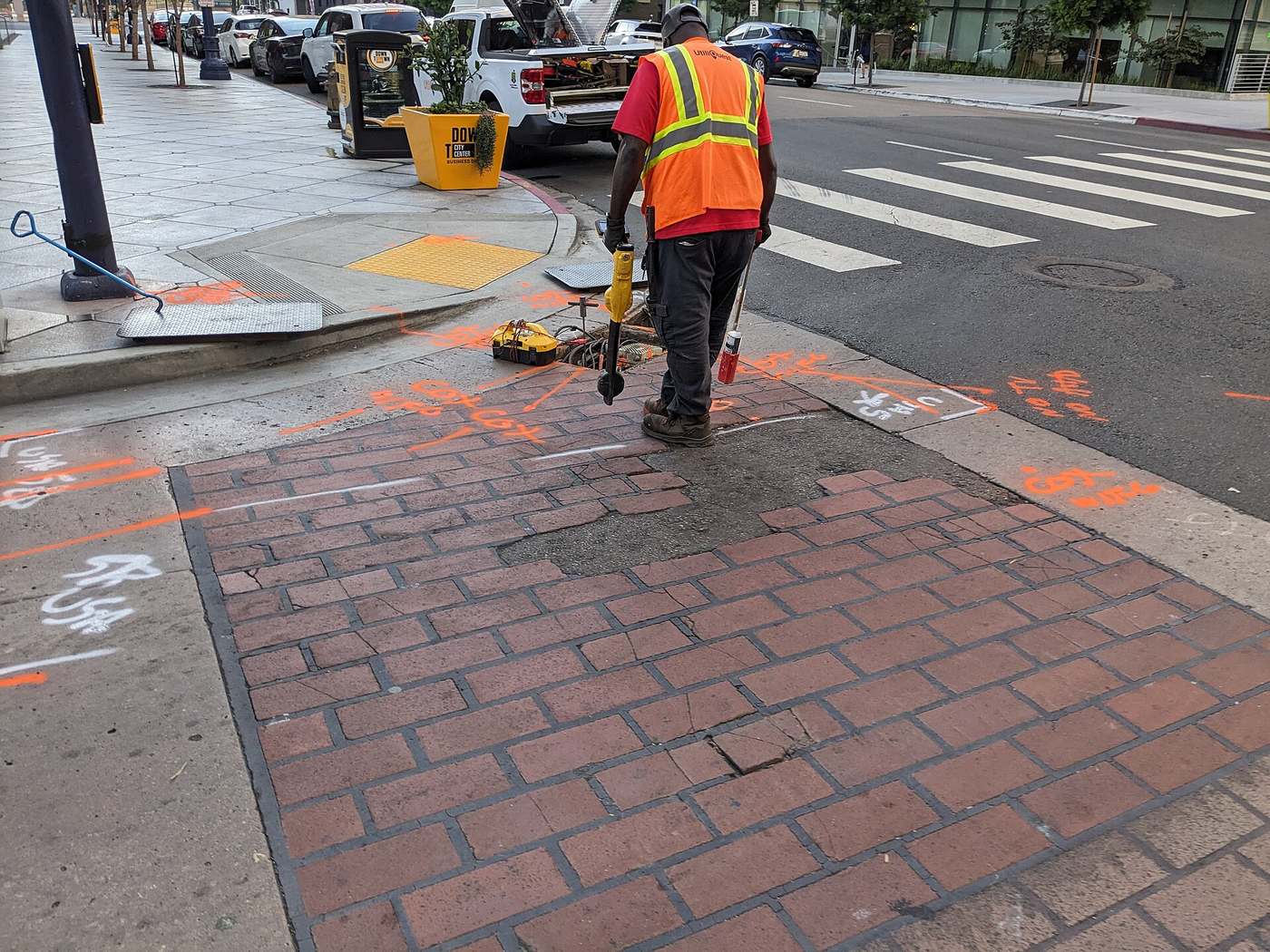 A locator in an orange vest walks a city sidewalk with a marking wand; fresh orange locate paint runs across the brick pavers.