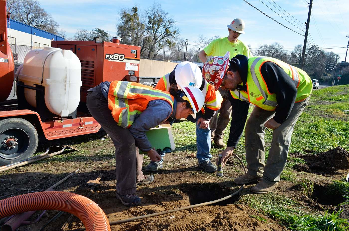 Two workers kneel over a fresh pothole in grass, looking down at a buried line, while others stand by a vacuum excavation unit.
